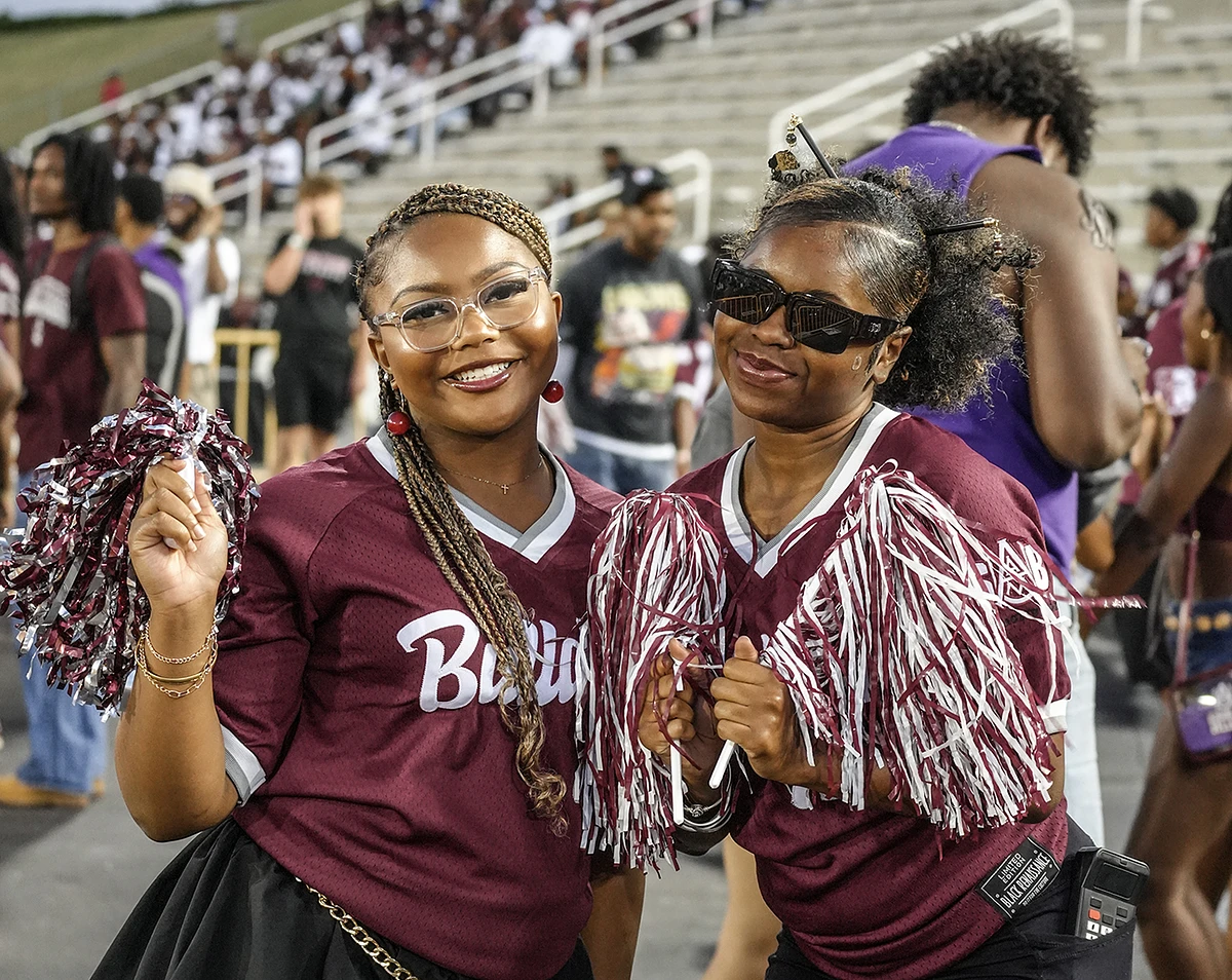 Two women in matching maroon jerseys, holding pom-poms, smile and pose for a photo at a crowded outdoor event.