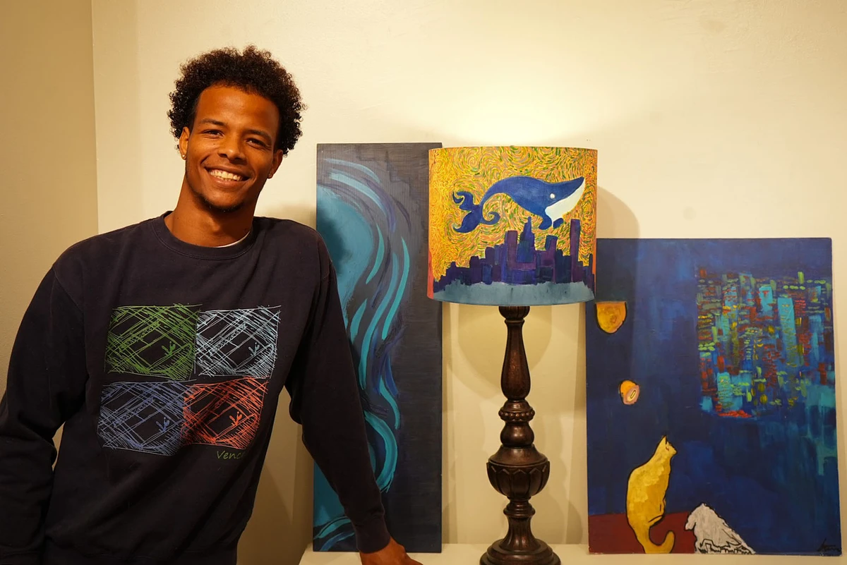 A smiling person stands next to colorful art pieces, including a lamp with a whale design.