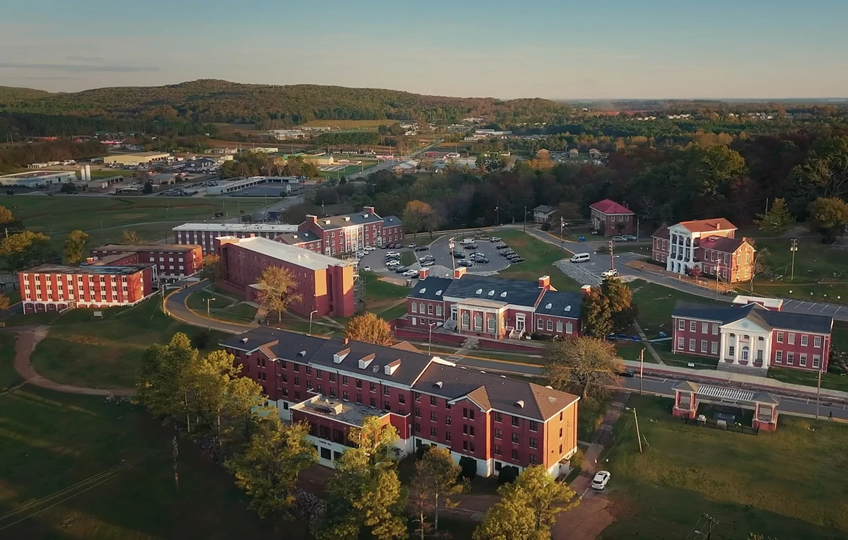 An aerial view of a campus with multiple red brick buildings, surrounded by trees and parking lots.