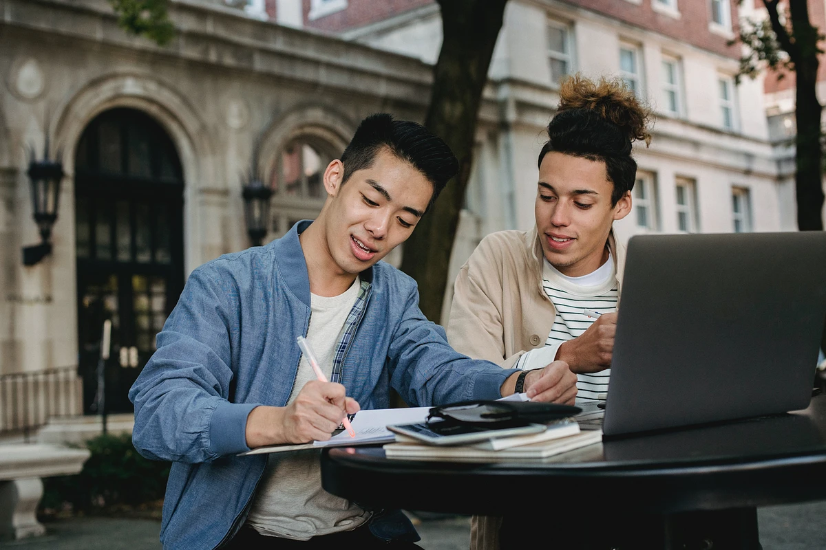 Two students sit at outdoor table, working on homework; surrounded by buildings and some trees